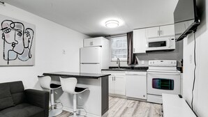 Een kleine keuken met witte kastjes, een magnetron, een fornuis en een koelkast. In de woonkamer staat een barkruk en een bank.