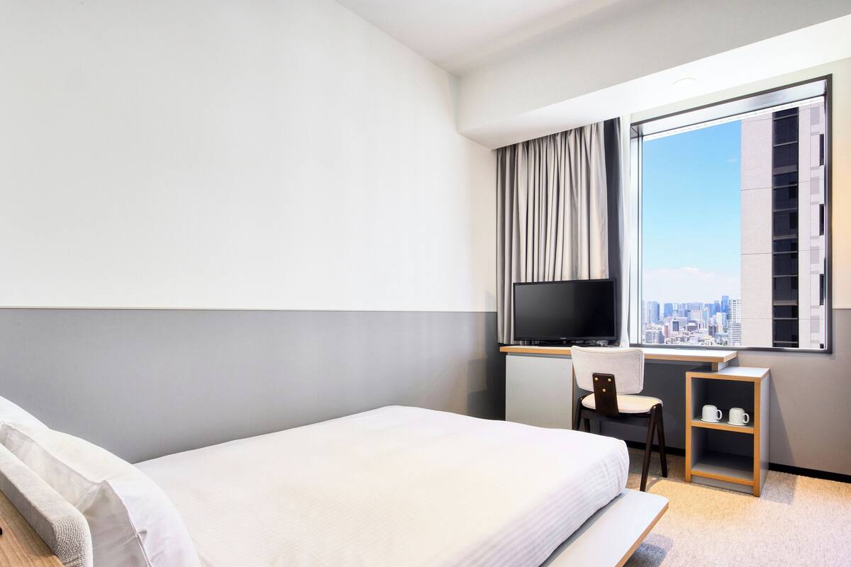 A hotel room with a bed, a desk, a chair, a TV, and a city view.