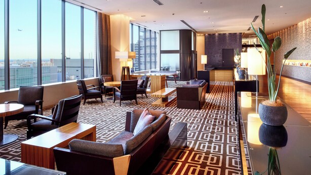 A modern hotel lobby with a high ceiling, large windows offering a city view, a seating area with sofas and chairs, and a reception desk. - LOISIR HOTEL SHINAGAWA SEASIDE (Tokyo)