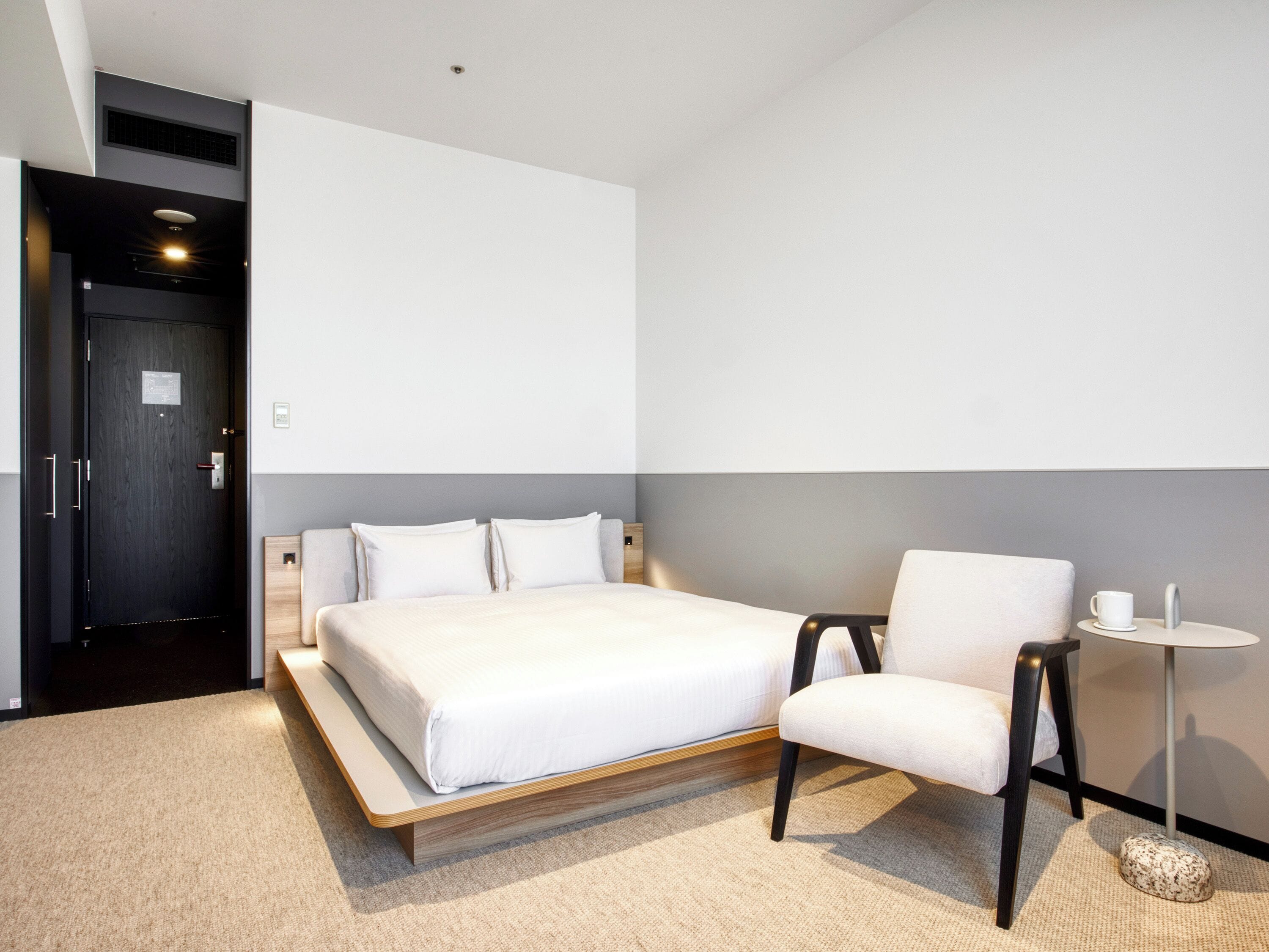 A modern hotel room with a large bed, a chair, a small table, and a wall-mounted heater.
