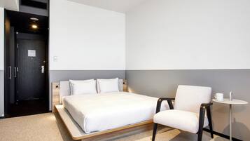 A modern hotel room with a large bed, a chair, a small table, and a wall-mounted heater.