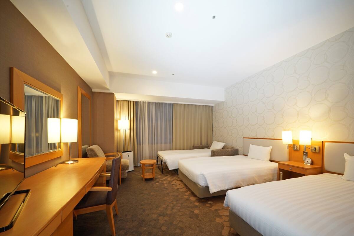 A hotel room with a desk, two beds, a chair, a small table, and a mirror.