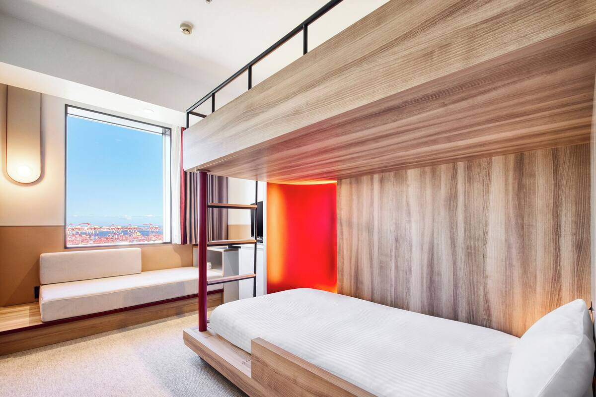 A modern hotel room with a bunk bed, a sofa, a view of the city, and a red accent wall.