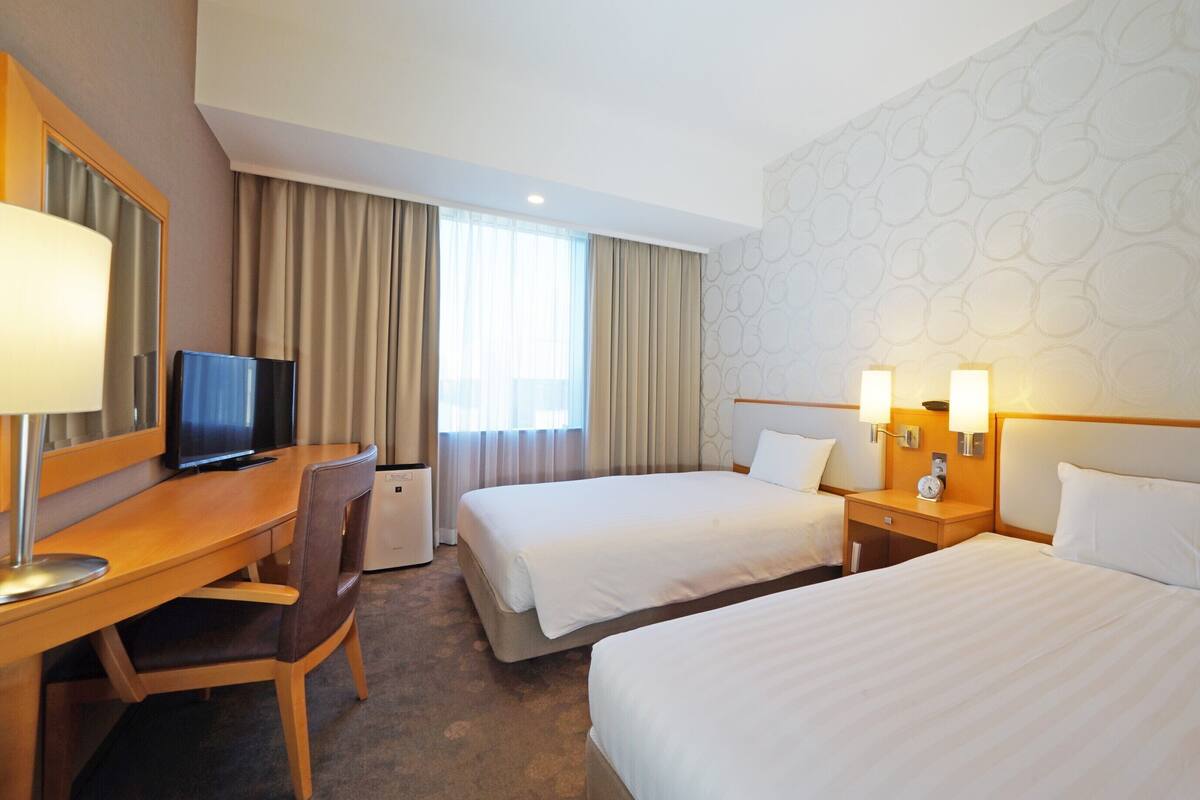 A hotel room with two beds, a desk with a chair, a television, and a large window with curtains.