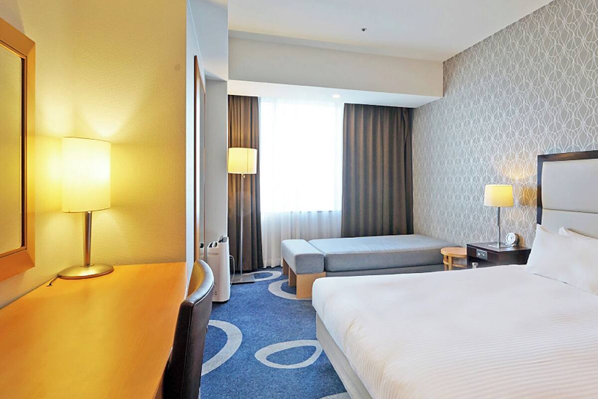 A hotel room with a large bed, a desk with a lamp, a bedside table, and a window with curtains.