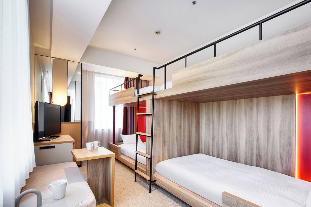 A modern hotel room with a bunk bed, a desk with a chair, a flat-screen TV, and a small seating area with a table and two chairs.