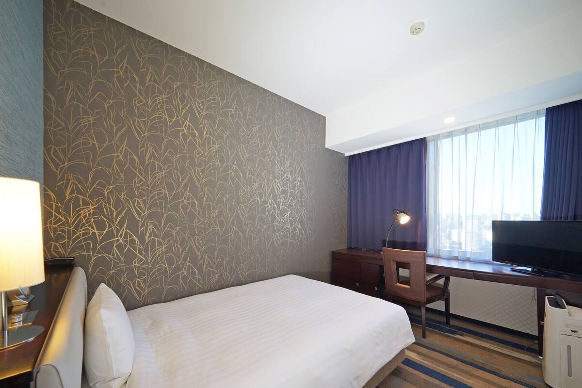 A hotel room with a bed, a desk with a computer, a TV, a lamp, and a window with curtains.