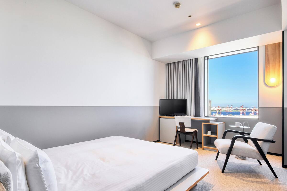 A modern hotel room with a bed, a desk, a TV, and a view of the city.