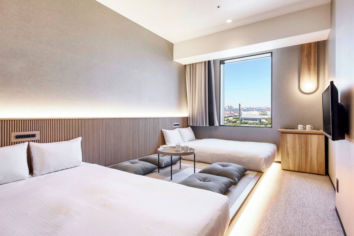 A modern hotel room with two beds, a small table, a TV, and a large window with a city view.