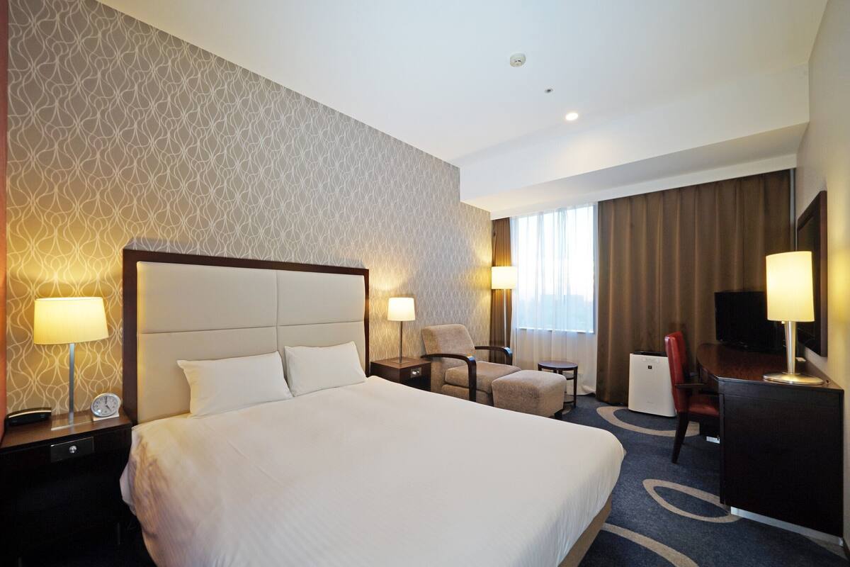 A hotel room with a large bed, a desk with a chair, a TV, and a window with curtains.