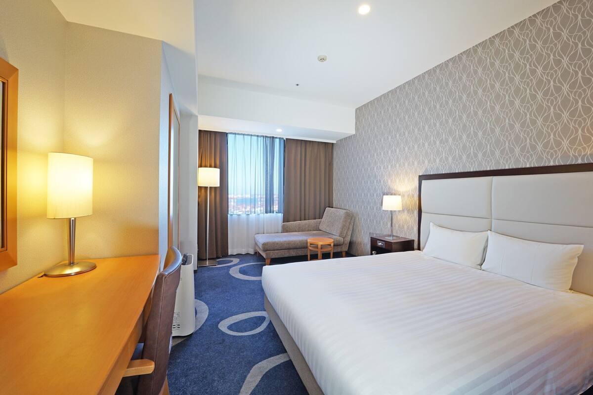 A hotel room with a large bed, a desk, a chair, a small table, and a window with curtains.