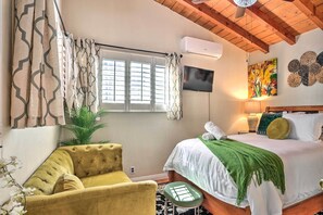 A bedroom with a bed, a yellow armchair, a green stool, a potted plant, and a window with curtains. - 1 Mi to Lake Gregory: Boho Crestline Cabin (Crestline)