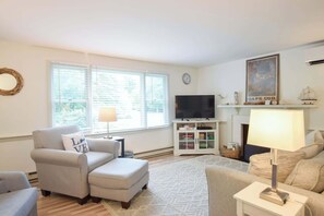 A living room with a sofa, ottoman, TV, fireplace, and a large window with blinds.