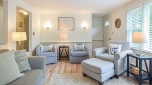 A living room with a sofa, two armchairs, a coffee table, and a rug. There are two wall-mounted lamps, a framed map, and a mirror on the wall. - Outdoor Oasis w Fire Pit, Hammock, 1 Mi to Beach! (Brewster)