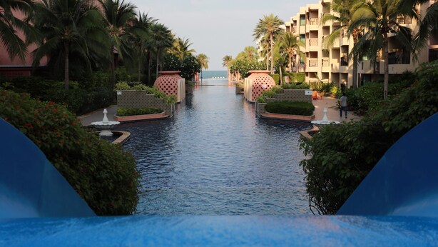 Área de piscina em resort, com palmeiras e vista para o oceano.