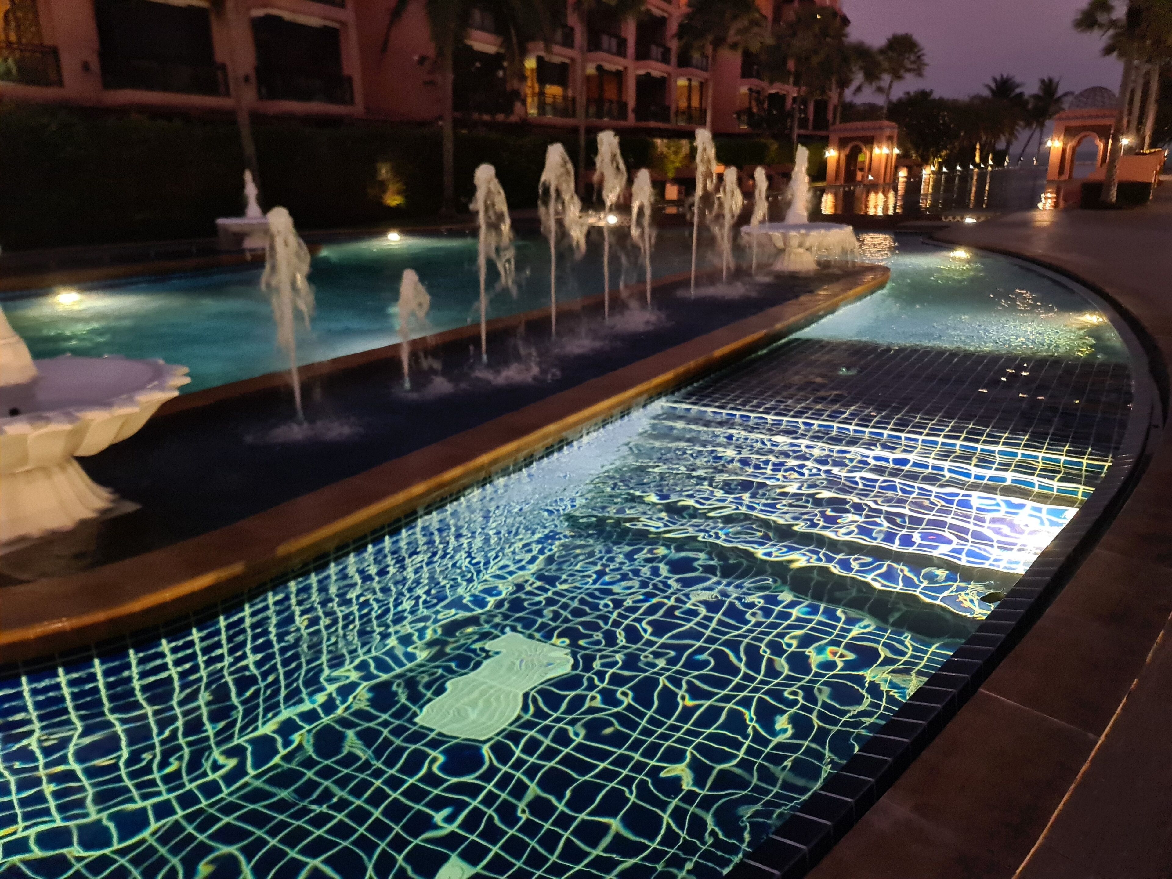 A nighttime swimming pool with water fountains and mosaic tiles.