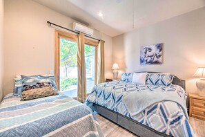 A bedroom with two beds, a window with curtains, a wall-mounted air conditioner, a bedside lamp, and a painting.