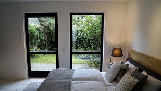 Ein Schlafzimmer mit Bett, Kissen, einem Nachttisch, einer Lampe und Blick auf einen Garten durch eine Schiebetür.