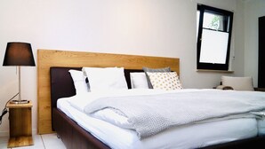 Ein ordentlich bezogenes Bett mit einem Holzkopfstück, einer Nachttischlampe und einem Fenster mit Jalousien.