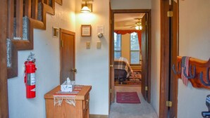A hallway with a wooden staircase, a fire extinguisher, a small desk with a tissue box, and a door leading to a bedroom with a bed and a window. - The Nestled Inn - Unit C3 (Ruidoso)