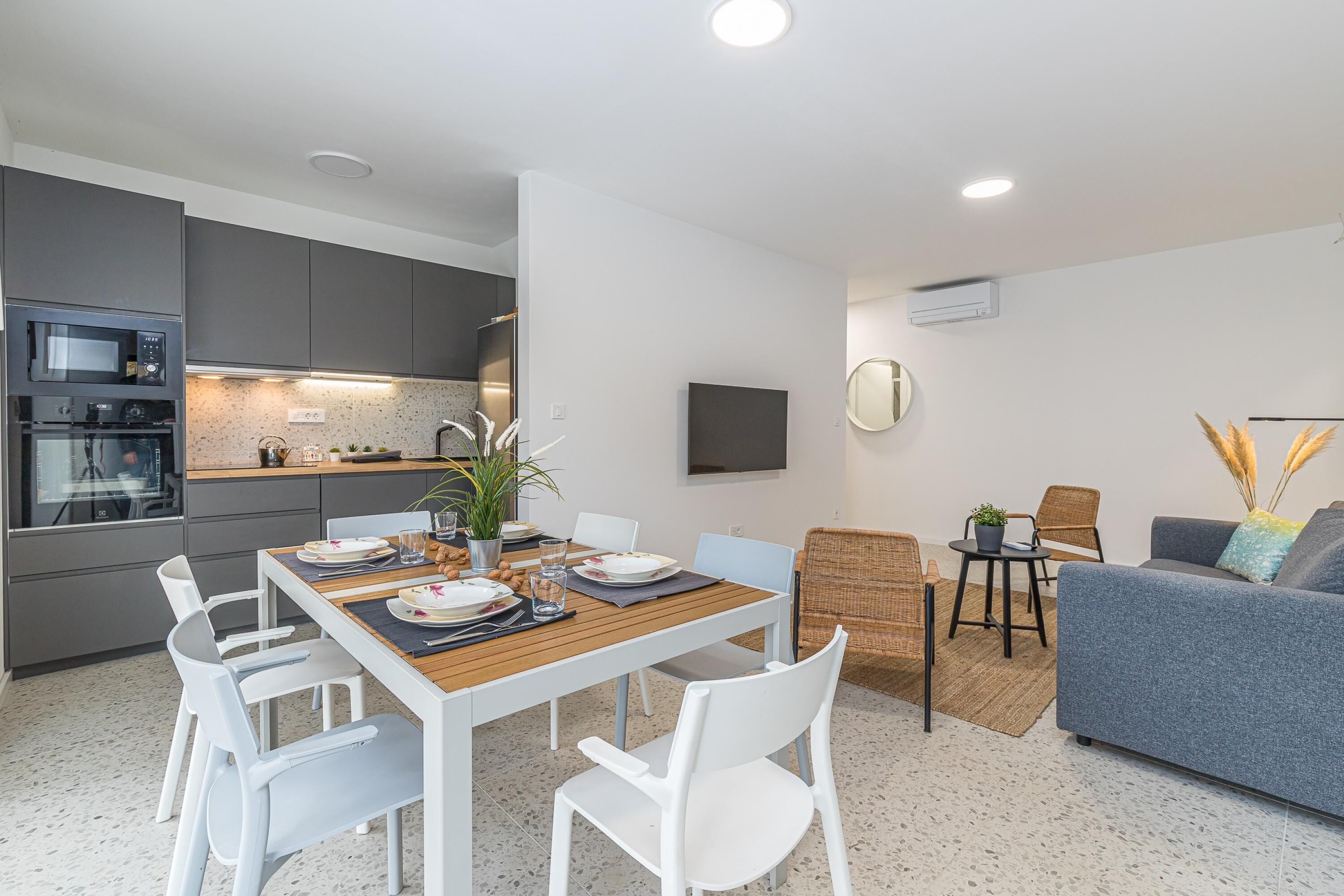 Una cocina moderna con electrodomésticos integrados, un comedor con mesa de madera y sillas blancas, un sofá gris y un televisor de pantalla plana montado en la pared.