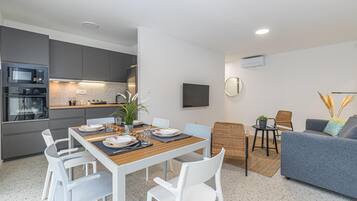Una cocina moderna con electrodomésticos integrados, un comedor con mesa de madera y sillas blancas, un sofá gris y un televisor de pantalla plana montado en la pared.