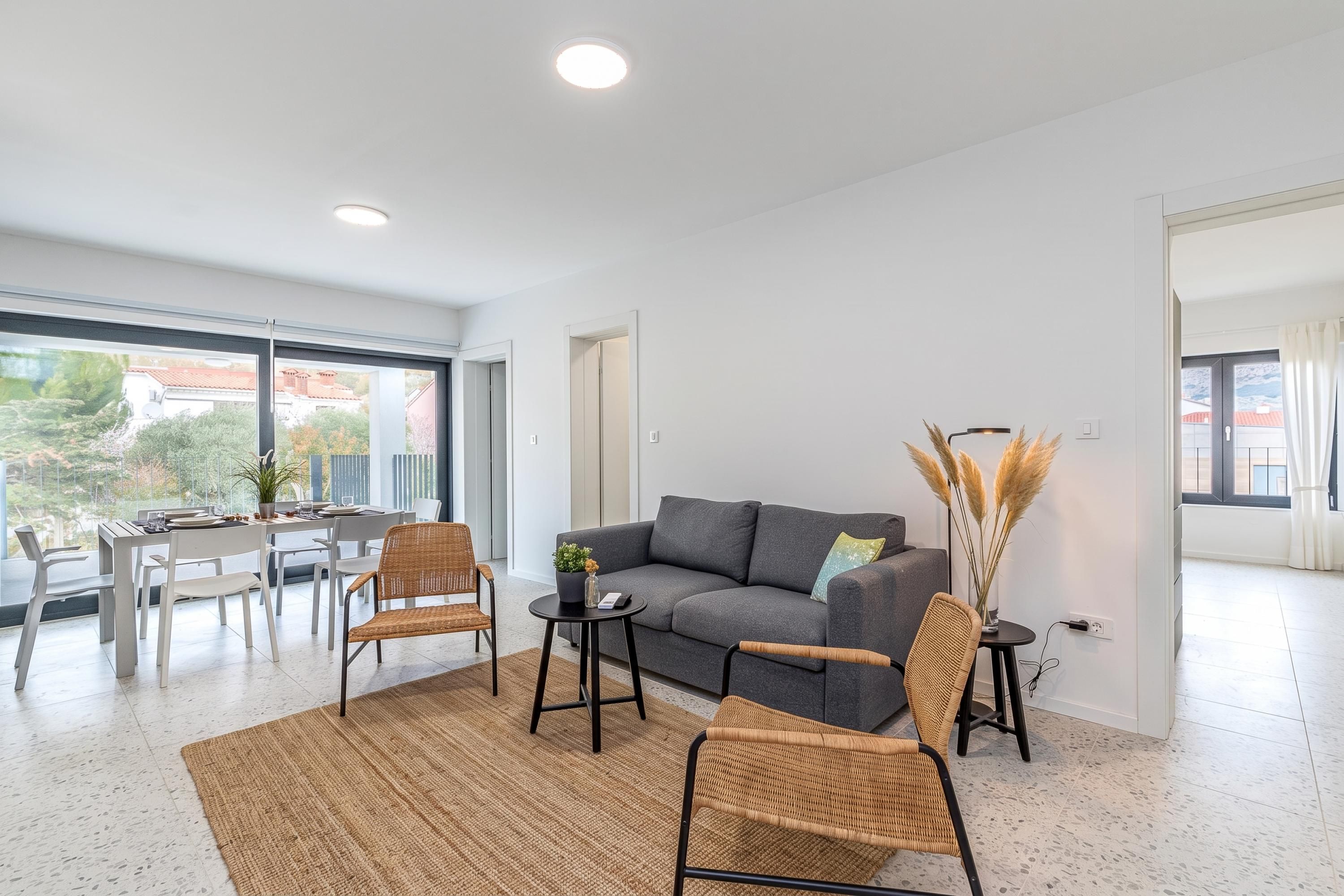A modern living room with a grey sofa, two wicker chairs, a small round table, and a large window with a view of buildings and trees.