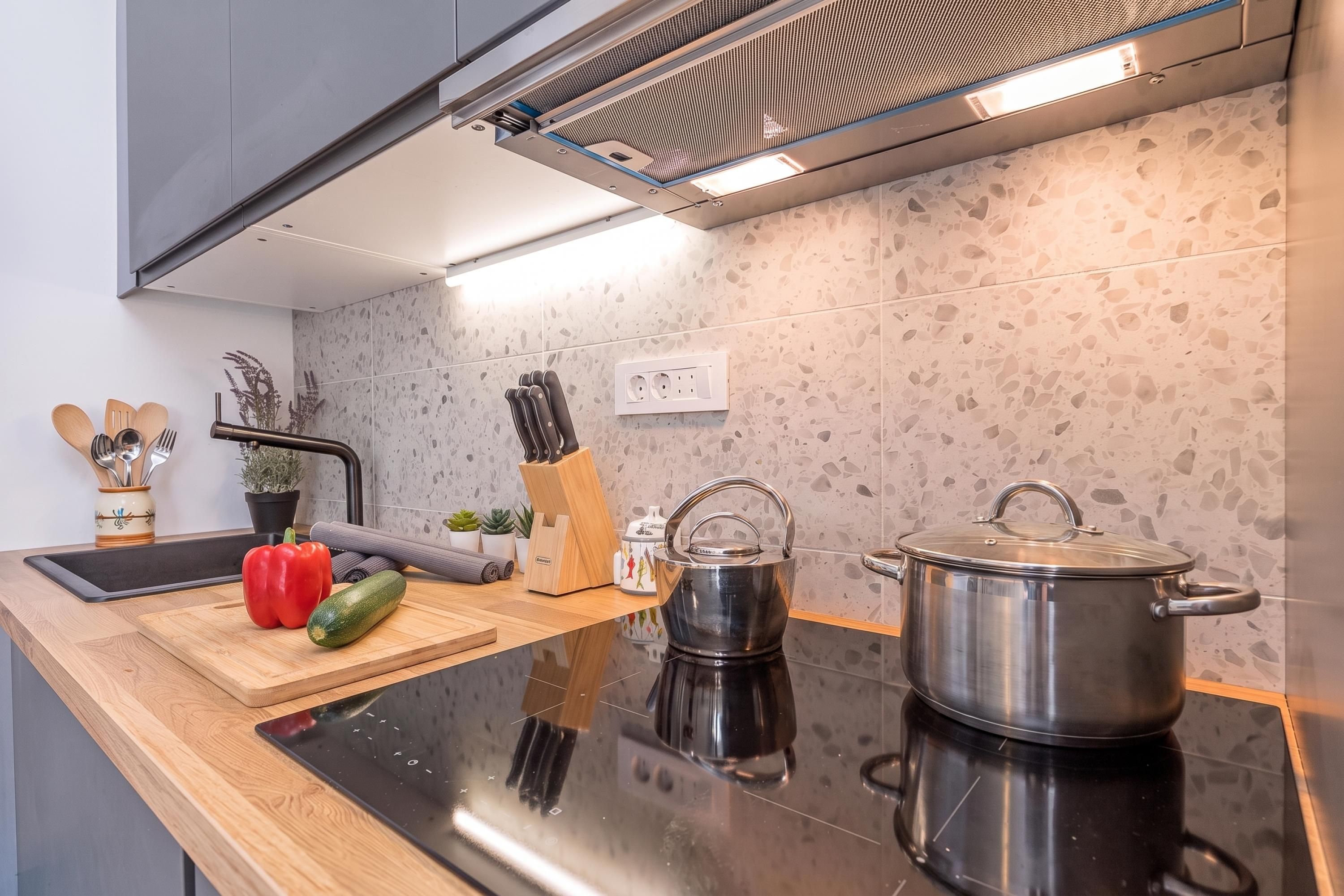 A modern kitchen with a black countertop, a stainless steel kettle, a pot, a cutting board with vegetables, and a knife block.