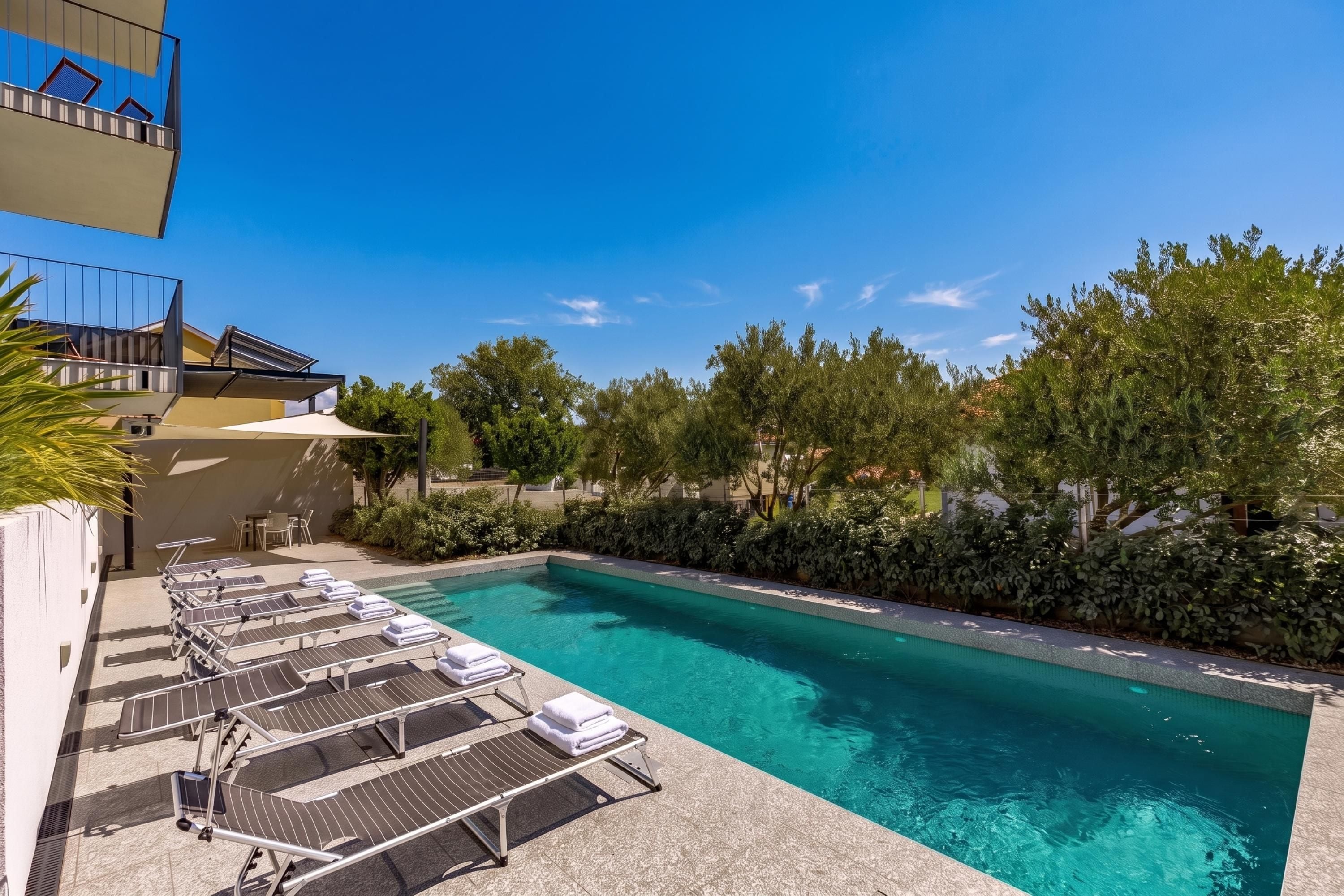 Una piscina con sillones de descanso y sombrillas, rodeada de vegetación y un cielo despejado.