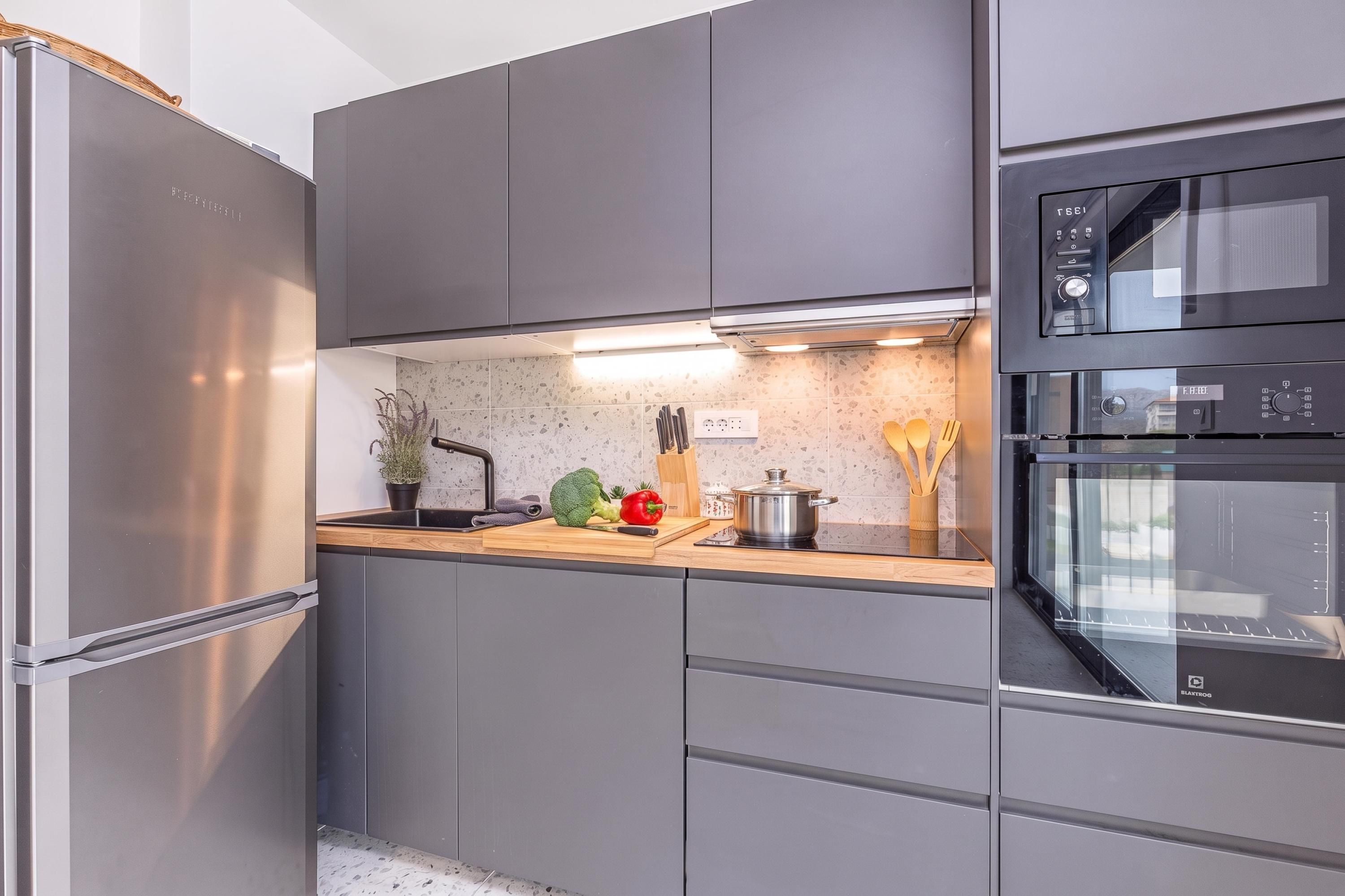 A modern kitchen with grey cabinetry, a stainless steel refrigerator, a built-in oven, and a countertop with a plant, vegetables, and utensils.