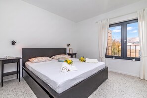 A modern bedroom with a large bed, bedside tables, a nightstand, a window with curtains, and a view of the outdoors. - Pool Sapore Di Mare Apartments (Baska)