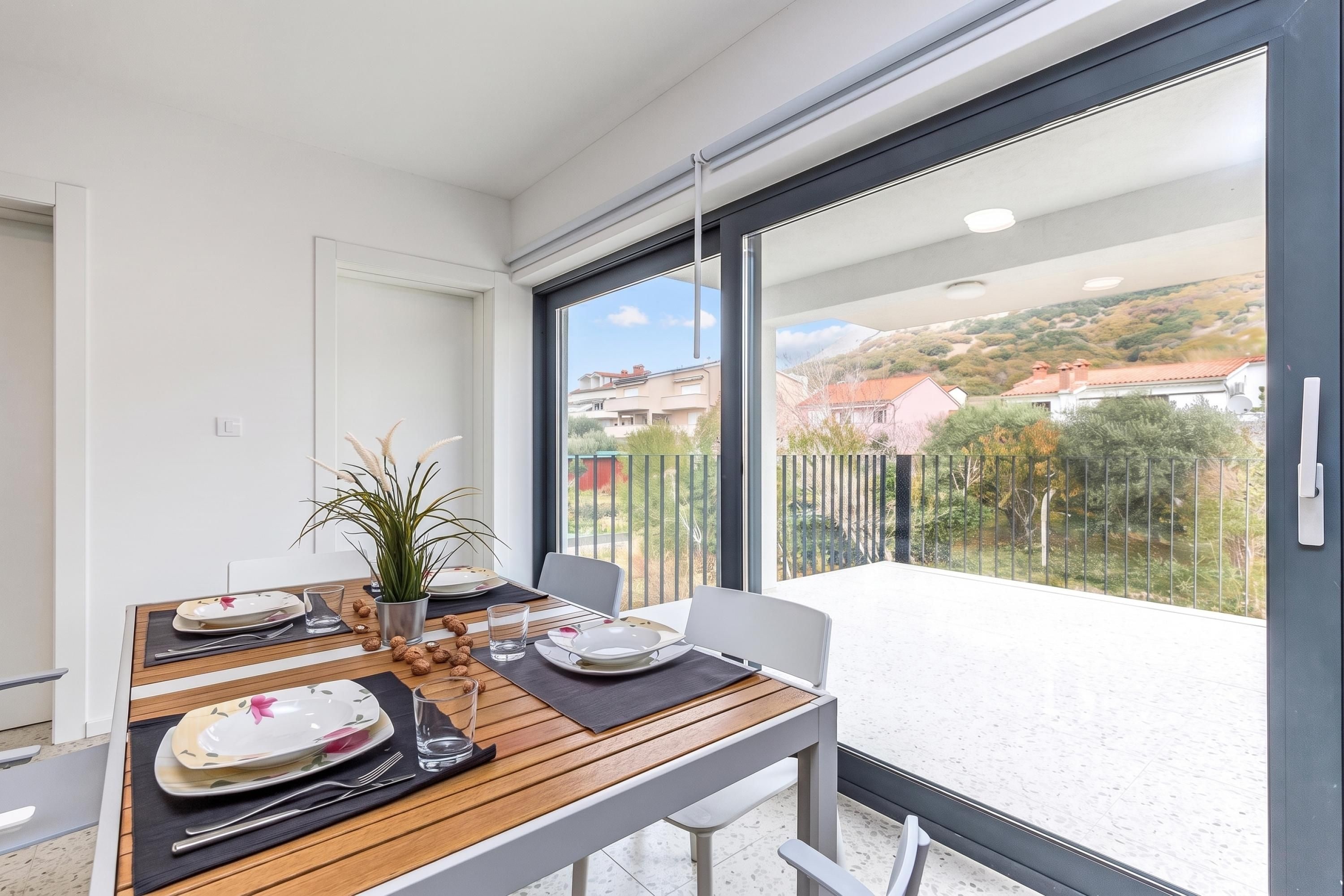 Un comedor con una mesa dispuesta para dos personas, una planta en maceta y vistas a un barrio residencial a través de grandes ventanales.