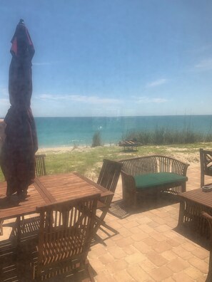 An outdoor seating area with wooden tables and chairs, overlooking a beach with clear blue water and a sandy shore.