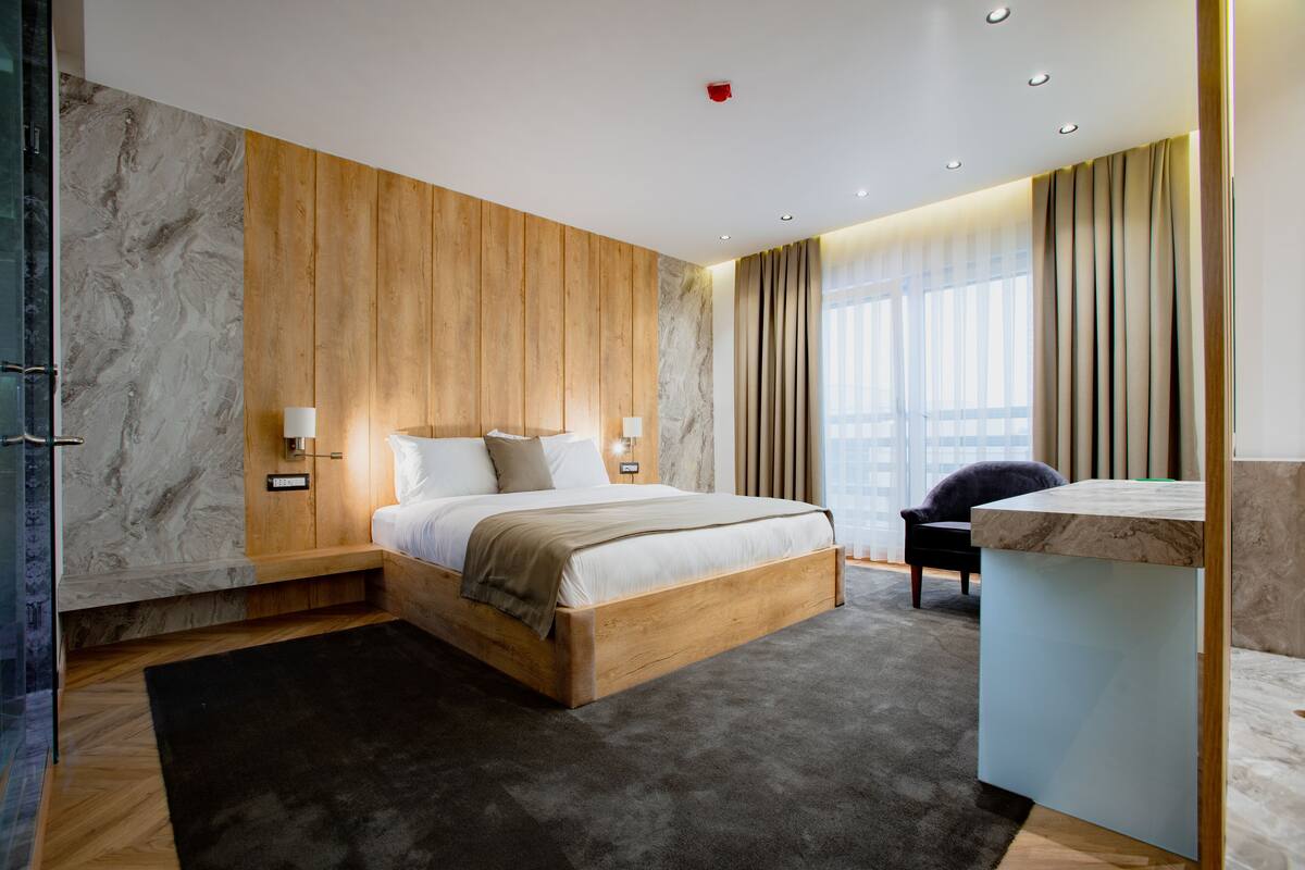 A modern hotel room with a wooden bed, a marble wall, a large window with curtains, and a small desk.