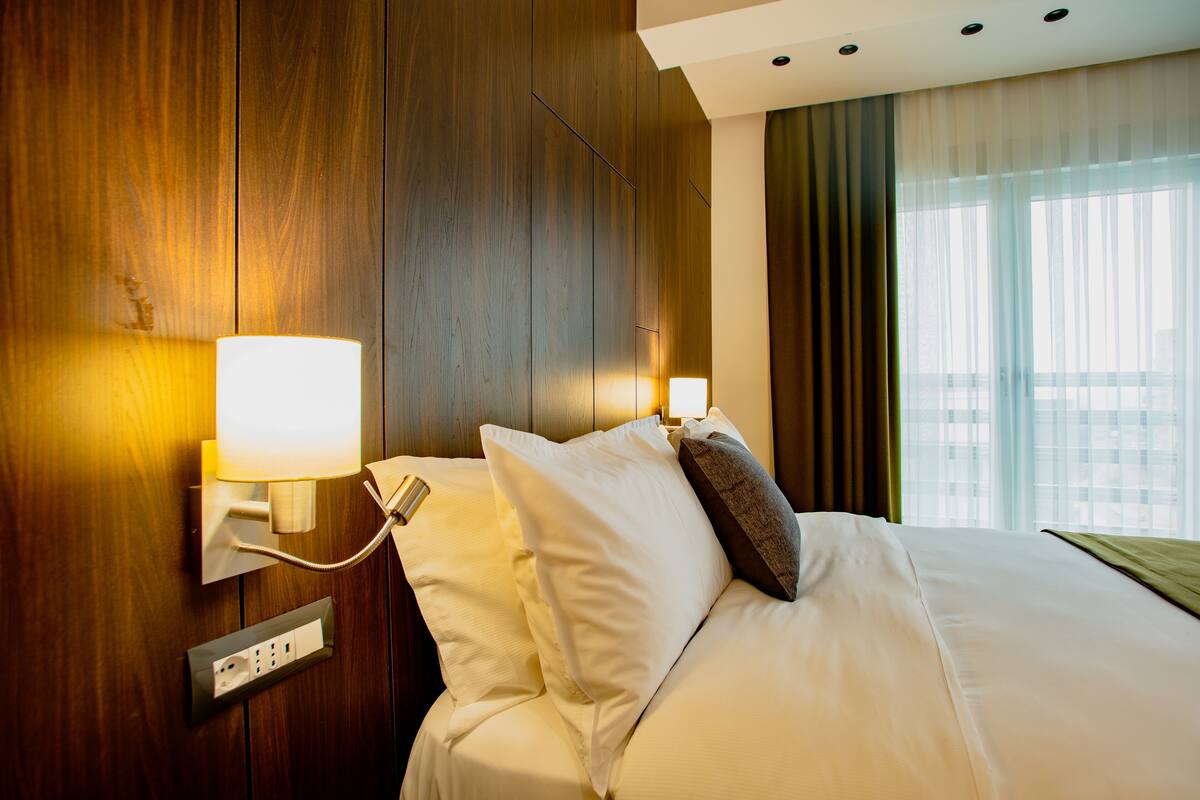 A hotel room with a wooden headboard, a bed with white linens, a bedside lamp, and a window with curtains.