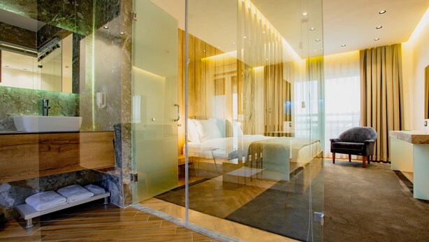 A modern hotel room with a glass partition, a wooden floor, a bed, a chair, and a bathroom with a marble countertop. - Ador Hotel (Pristina)