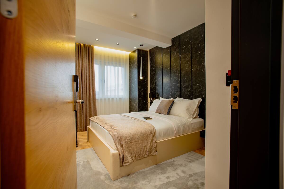 A modern bedroom with a large bed, a bedside table, a lamp, and a window with curtains.