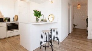Une cuisine moderne avec des meubles blancs, un four encastré, deux tabourets de bar et un petit îlot central agrémenté d’un vase de fleurs.