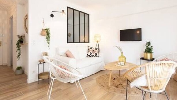 Un salon moderne avec un canapé blanc, des fauteuils en osier, une petite table ronde et un téléviseur à écran plat fixé au mur.