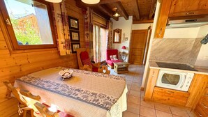 Een gezellig houten chalet met een eethoek, bestaande uit een tafel met een tafelkleed met dessin, houten stoelen en een mand. Een rode stoel en een kleine tafel met een lamp zijn ook te zien.