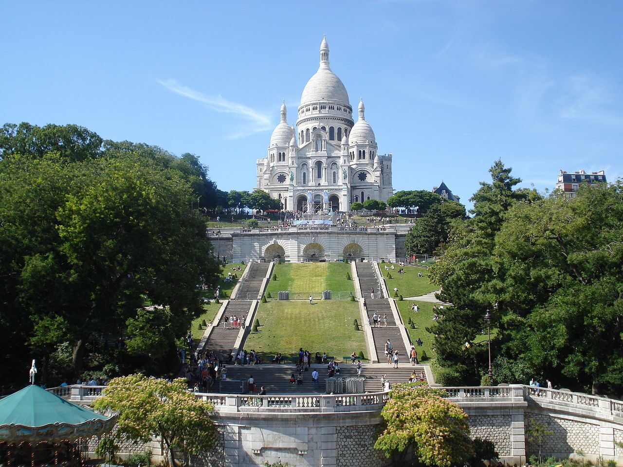 A large, white, domed building with multiple staircases leading up to it, surrounded by trees and a grassy area.