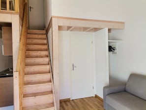 Una escalera de madera lleva a una puerta blanca, un sofá gris y un pequeño estante blanco con un cuadro.