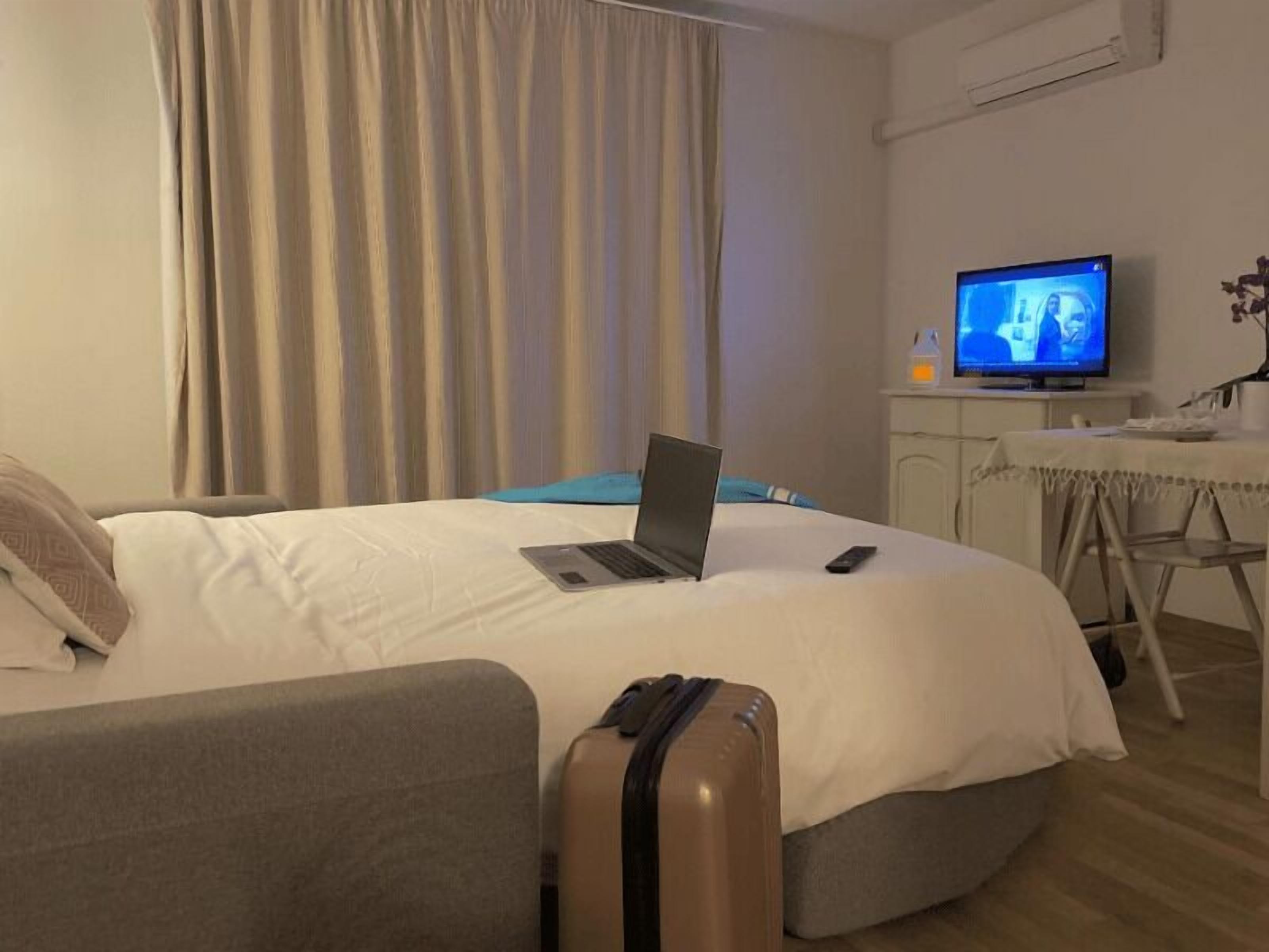 A hotel room with a bed, a laptop, a remote control, a suitcase, a television, and a small table with a chair.
