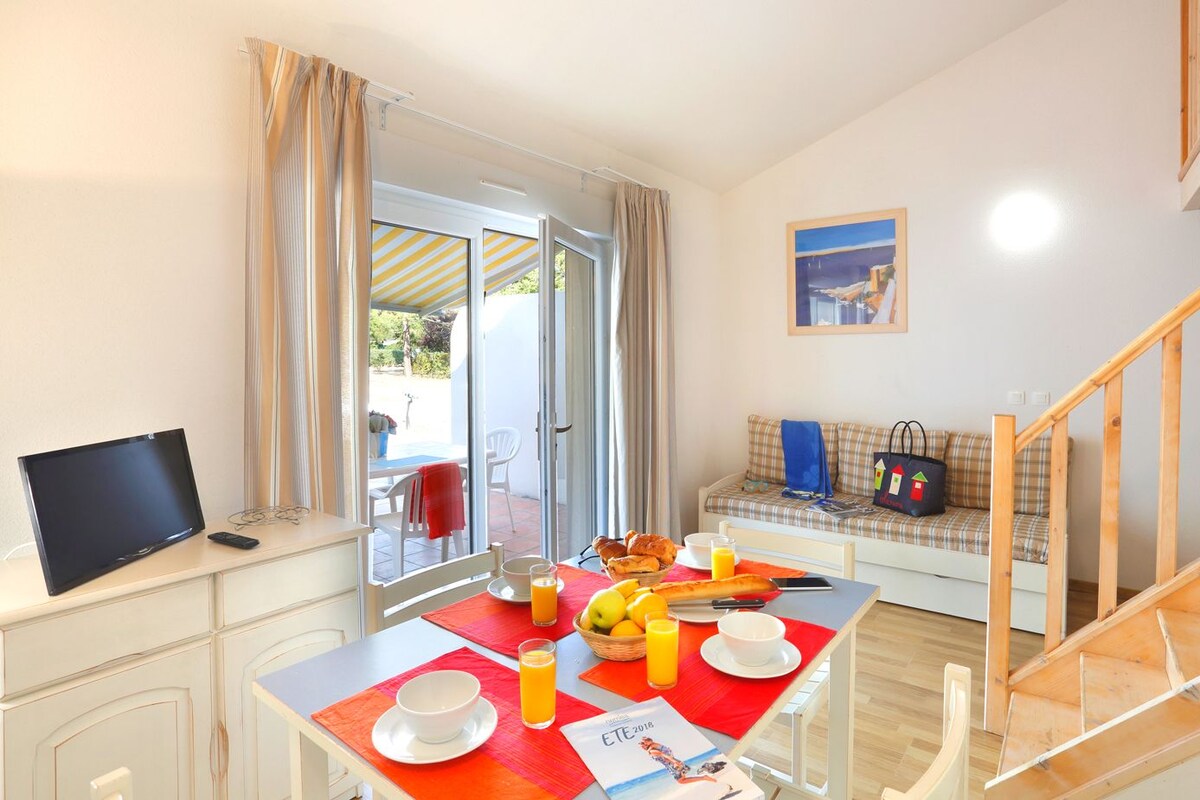 Un comedor con una mesa preparada para el desayuno, un televisor sobre un escritorio y un sofá con un bolso y un cuadro al fondo.
