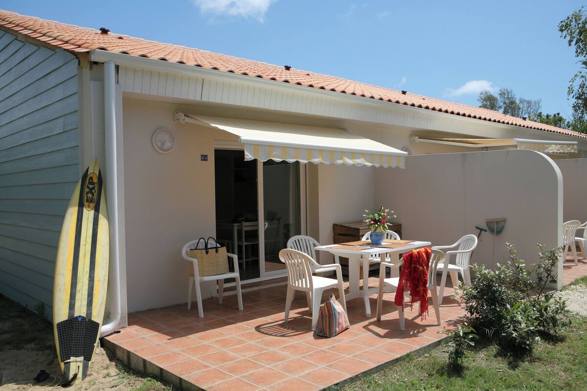 Eine überdachte Terrasse mit Tisch und Stühlen, ein Surfbrett, das an der Wand lehnt, und ein kleiner Gartenbereich.