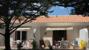 A building with a tiled roof, outdoor seating with chairs and tables, and a surfboard leaning against the wall. - Olydea Oléron les Sables Vigniers (Saint-Georges-d'Oleron)