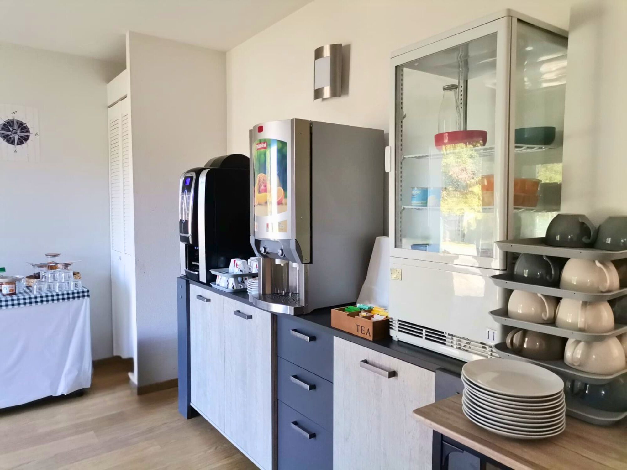 A kitchenette with a coffee machine, tea dispenser, and a glass cabinet with dishes.
