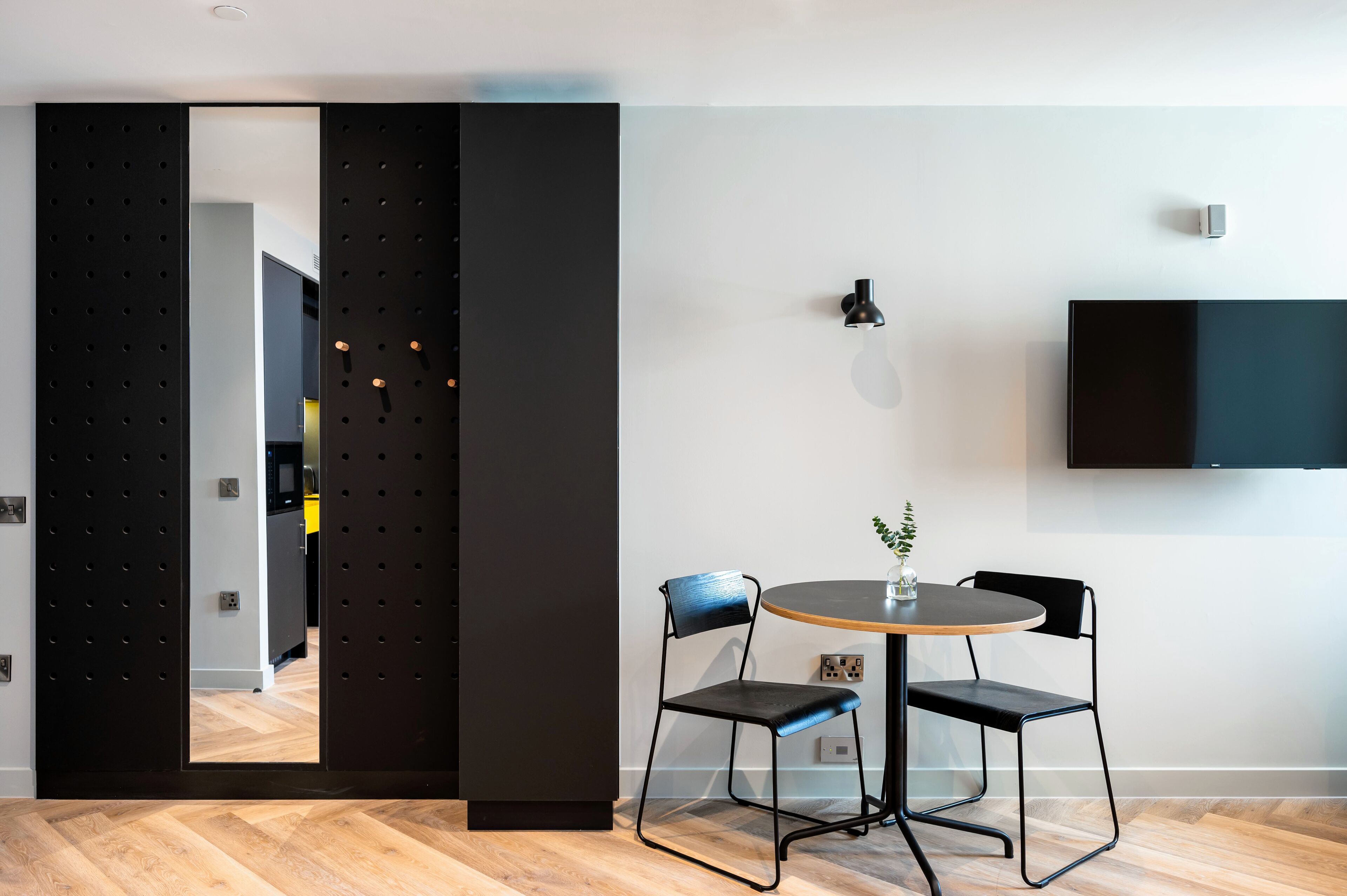 A modern room with a wooden floor, a round table with two chairs, a black-framed TV on the wall, and a sliding door with a perforated design.