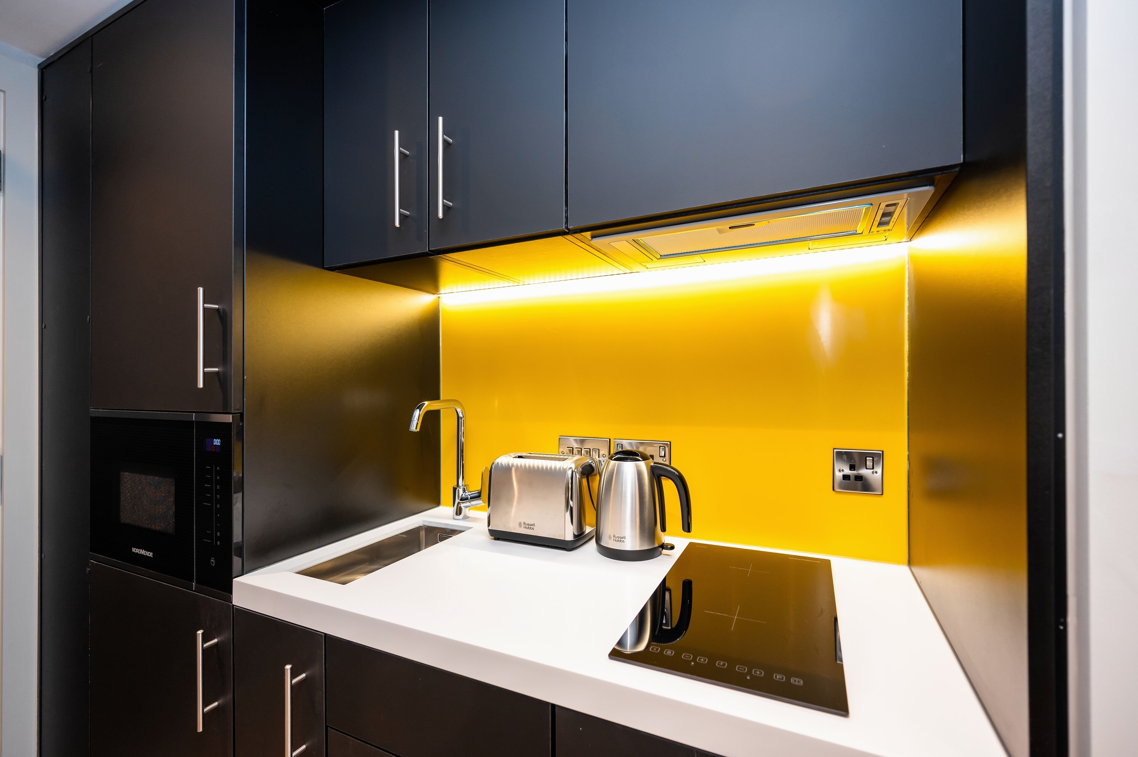 A modern kitchen with black cabinets, a white countertop, a built-in oven, a toaster, a kettle, and a sink.