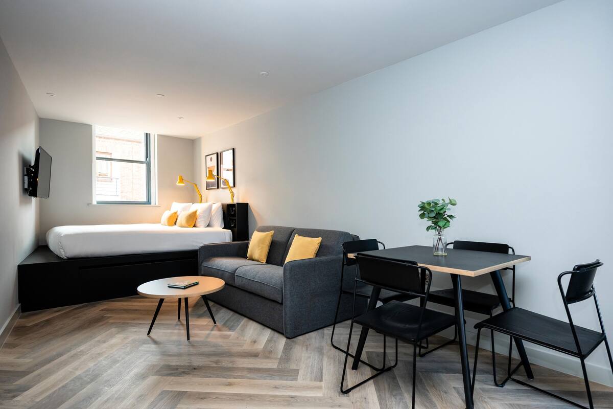 A modern, compact room with a bed, sofa, dining table, and chairs.